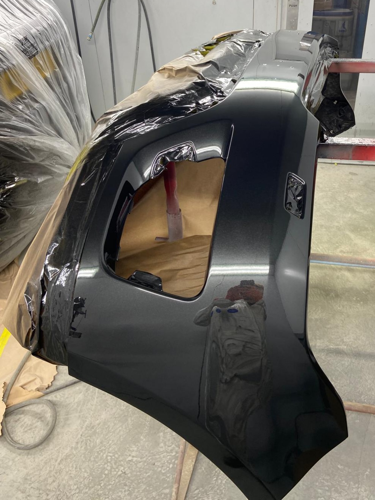 The bumper cover back from the paint booth in gloss black, still on the paint stand with the masking partly removed